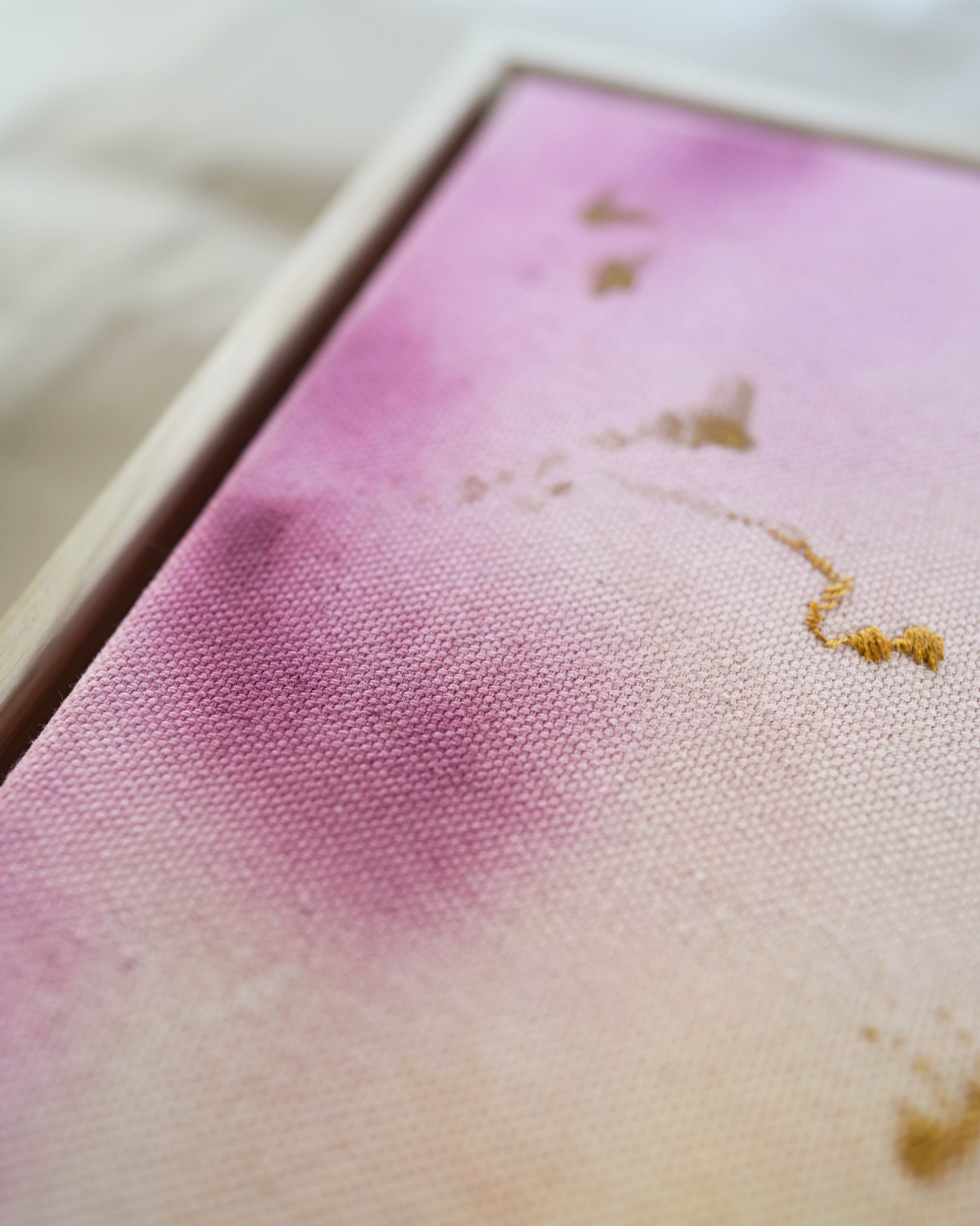 Abstract painting featuring soft pink, purple, and orange hues blending together with embroidery details of gold and dark purple on a canvas, framed in light wood.