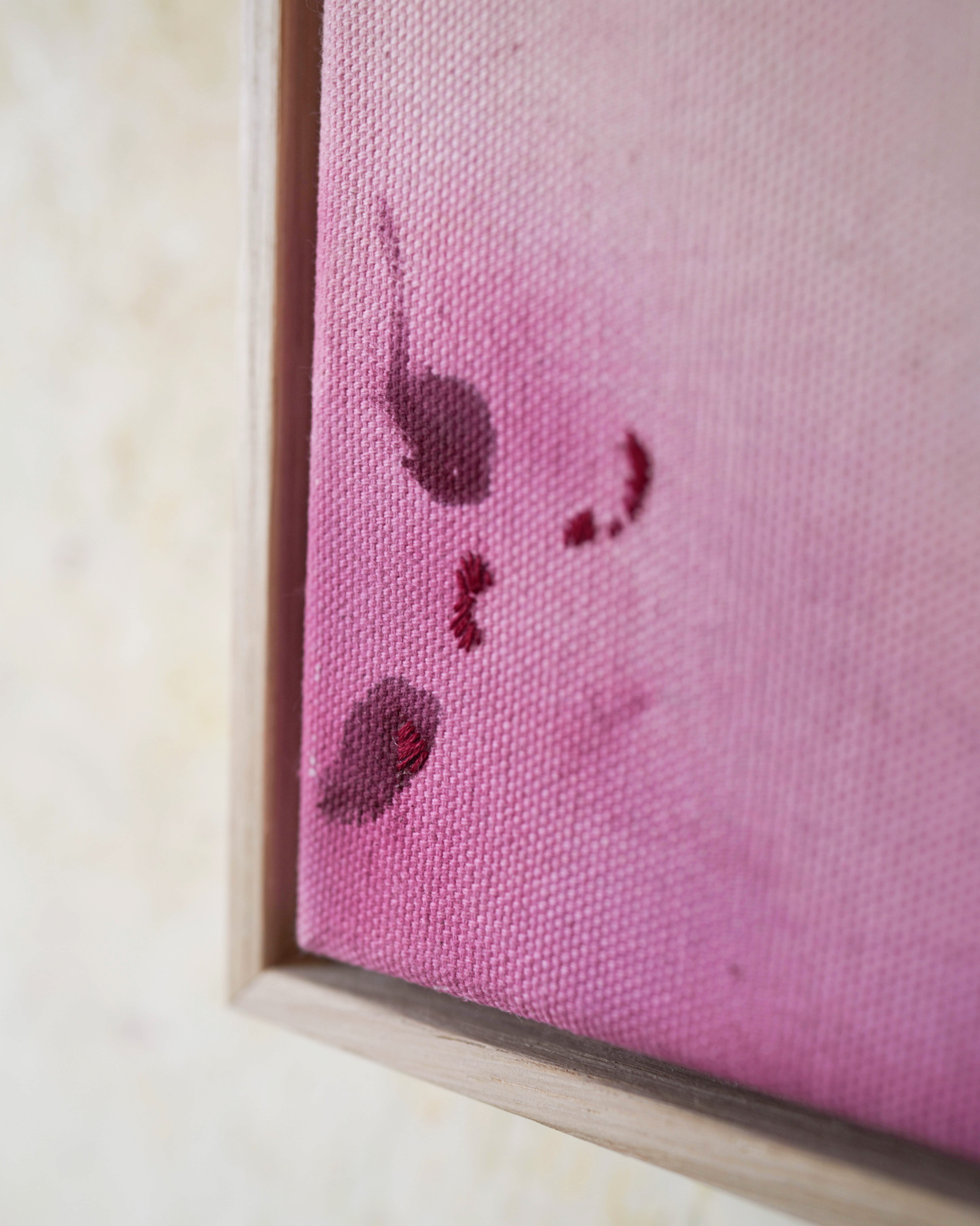 Abstract painting featuring soft pink, purple, and orange hues blending together with embroidery details of gold and dark purple on a canvas, framed in light wood.