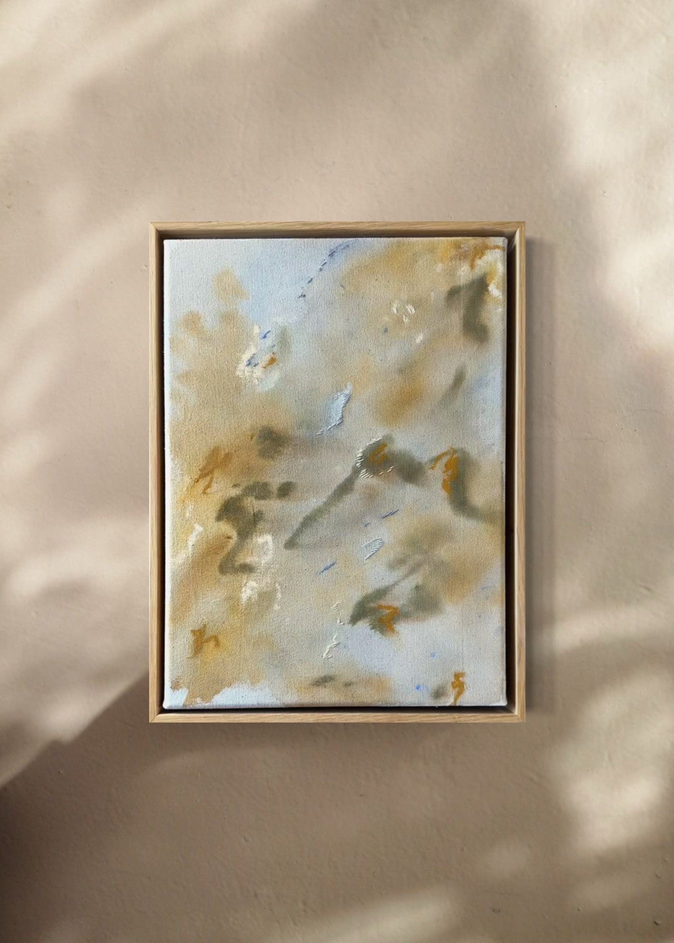 Abstract painting featuring soft, blended colors in beige, brown, and hints of blue. The artwork has a textured, stitched surface and is framed in a light oak wood frame.