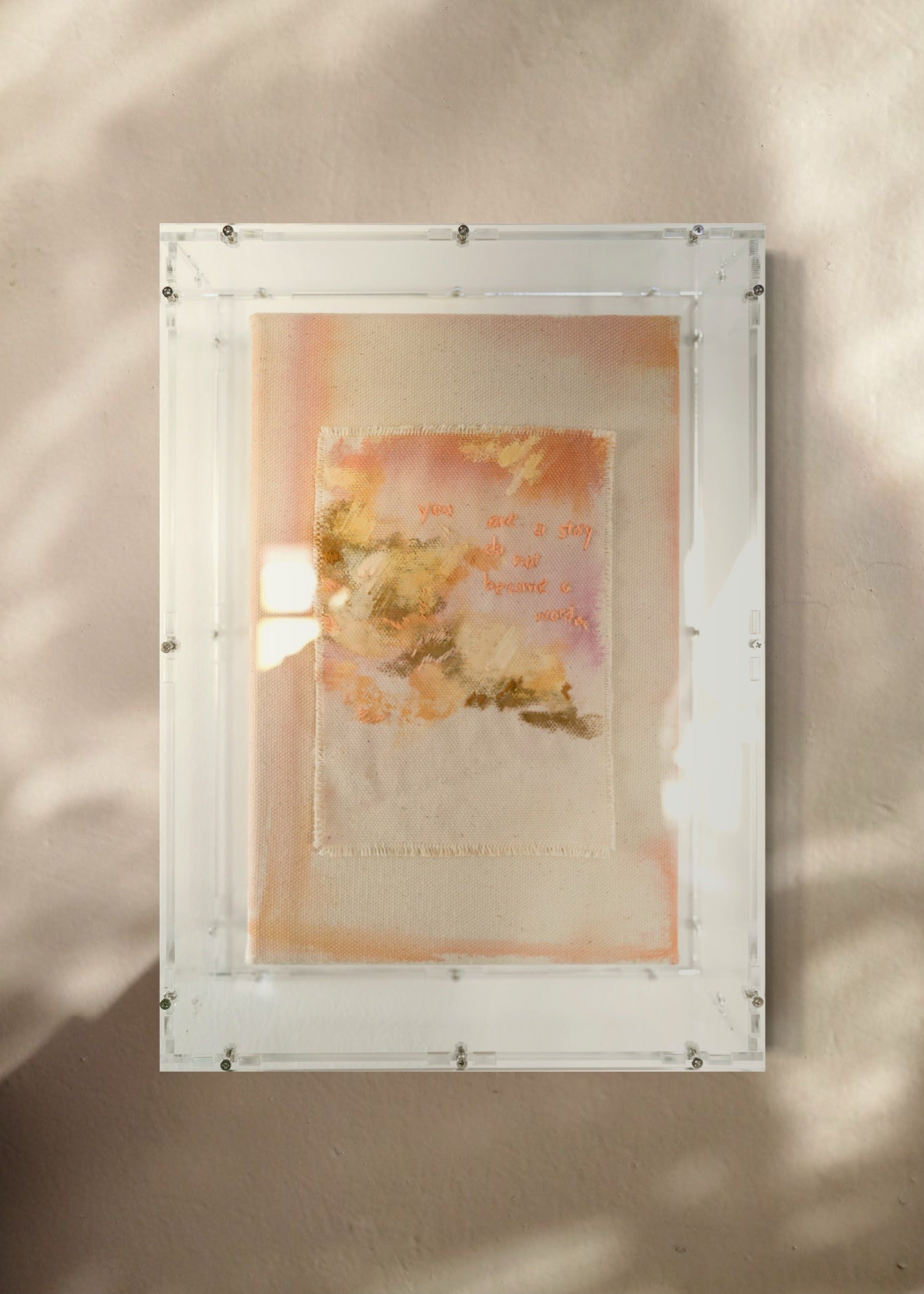 Textured abstract art with pastel hues of pink and orange, featuring delicate brushstrokes and embroidered text reading "you are a story do not become a word." Framed in clear acrylic.