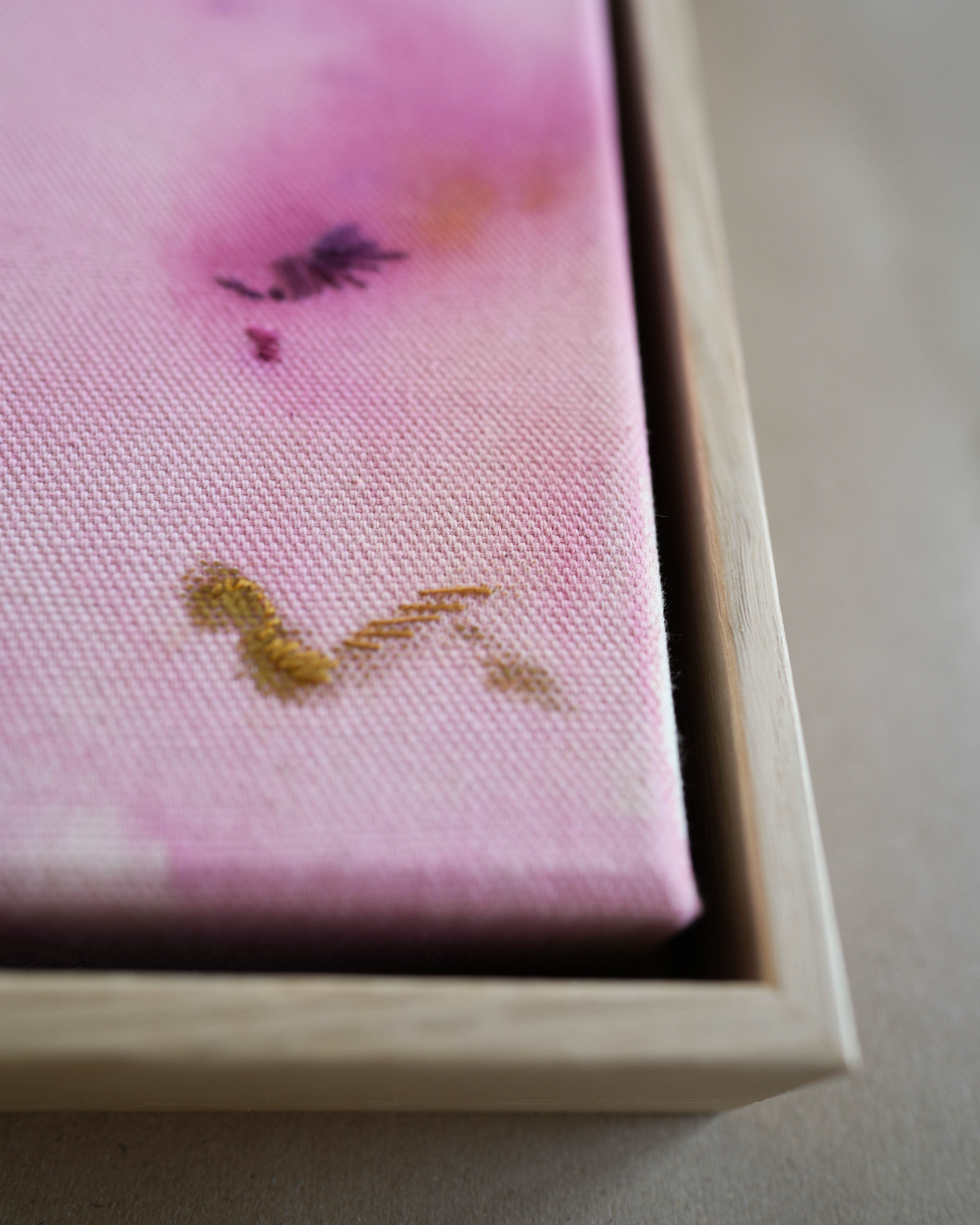Abstract painting with soft pink, purple, and orange colors blending together. It has gold and dark purple embroidery details on a canvas. The painting is framed in light wood.
