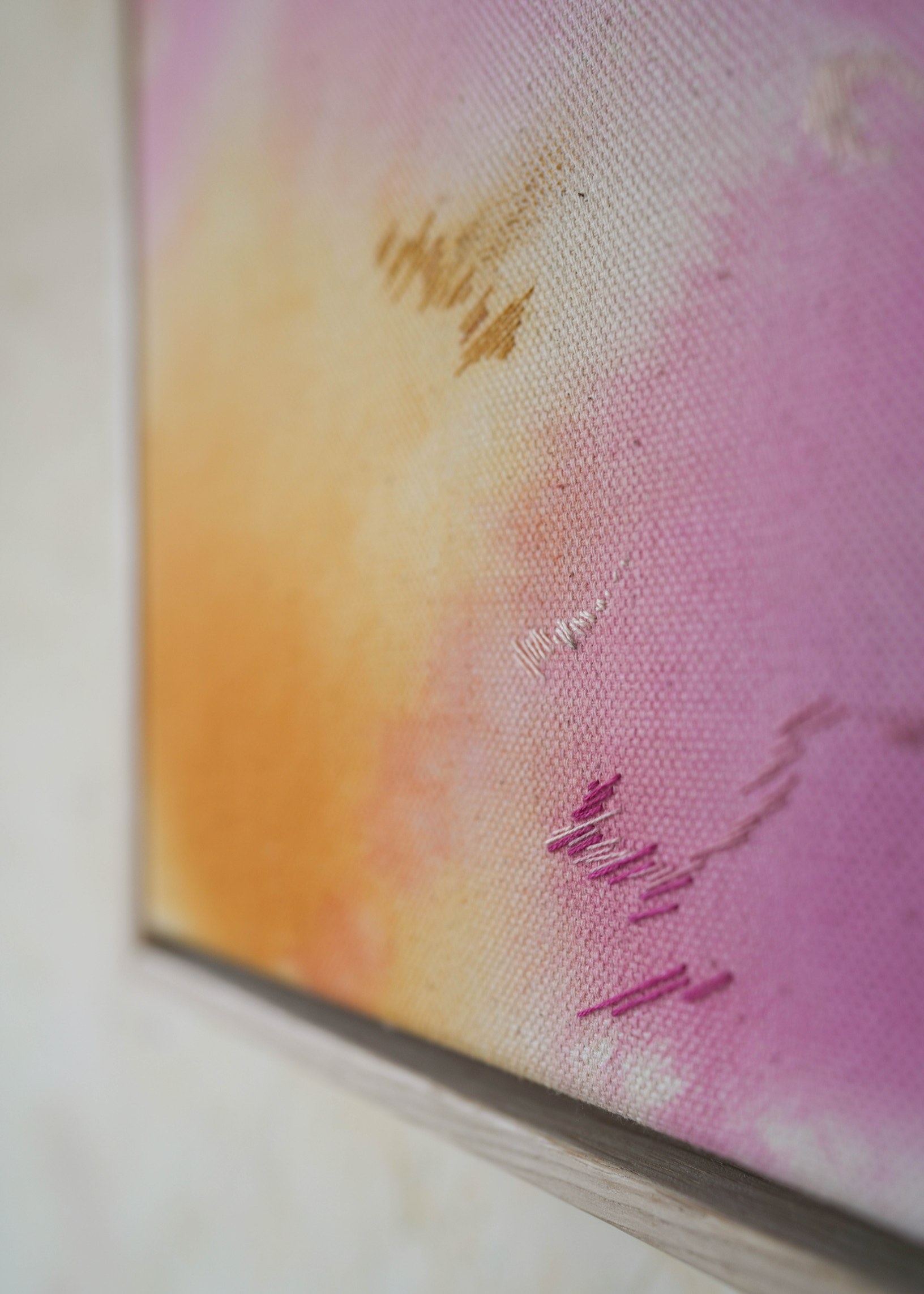 Abstract painting with soft pink, purple, and orange colors blending together. It has gold and dark purple embroidery details on a canvas. The painting is framed in light wood.