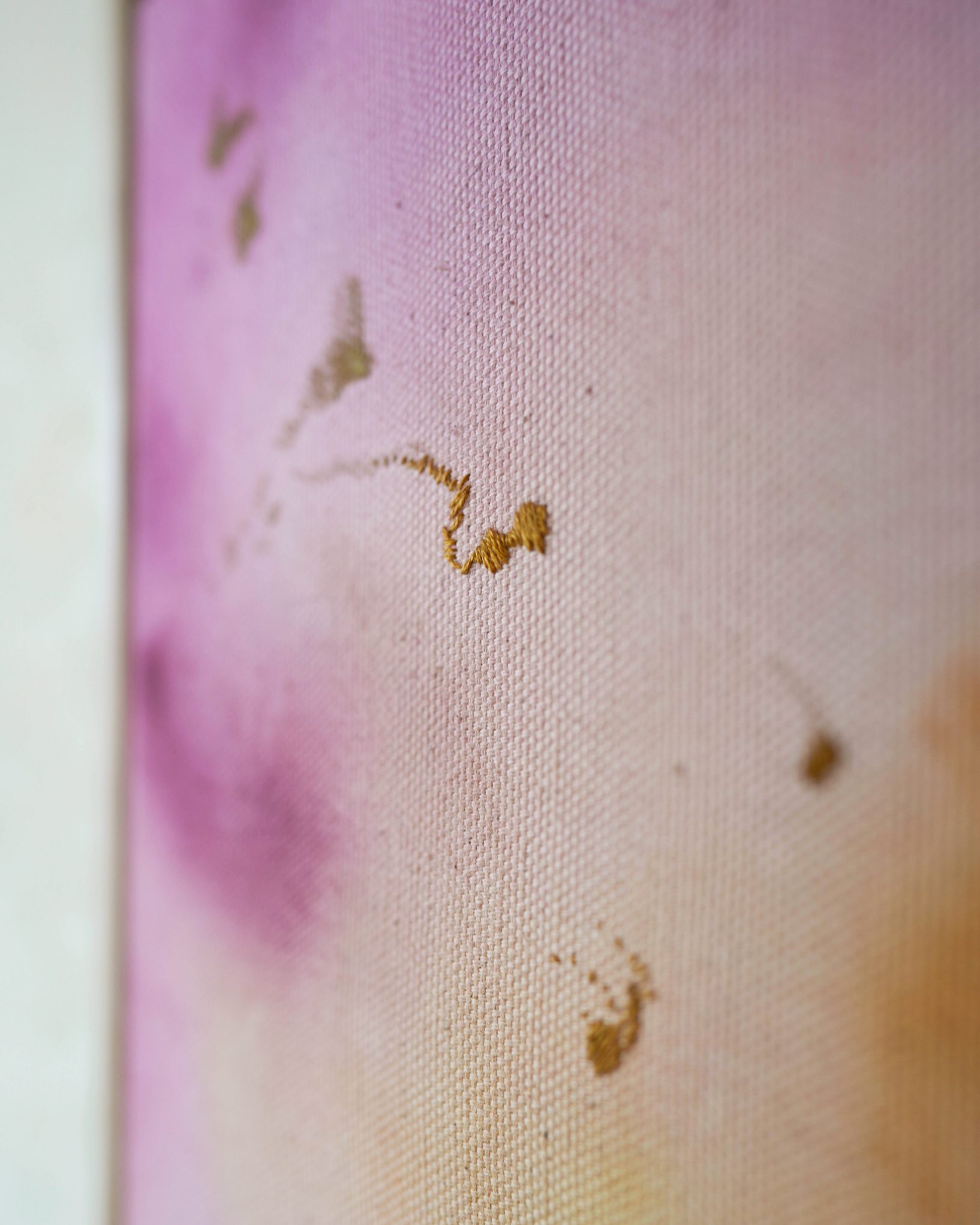 Abstract painting featuring soft pink, purple, and orange hues blending together with embroidery details of gold and dark purple on a canvas, framed in light wood.