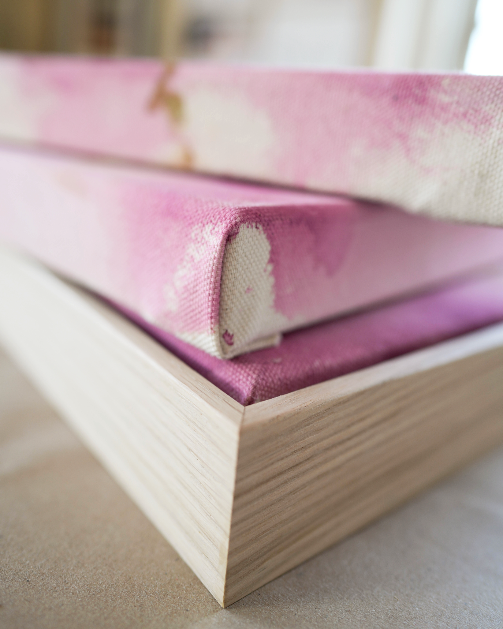 Abstract painting featuring soft pink, purple, and orange hues blending together with embroidery details of gold and dark purple on a canvas, framed in light wood.
