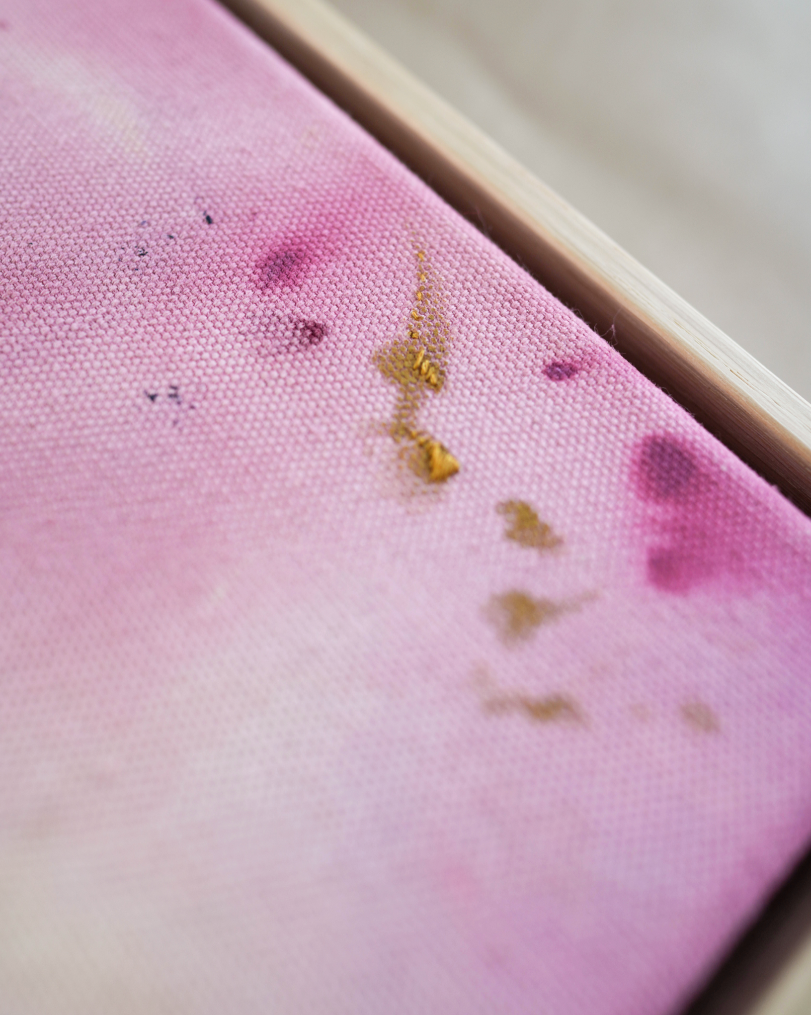 Abstract painting featuring soft pink, purple, and orange hues blending together with embroidery details of gold and dark purple on a canvas, framed in light wood.
